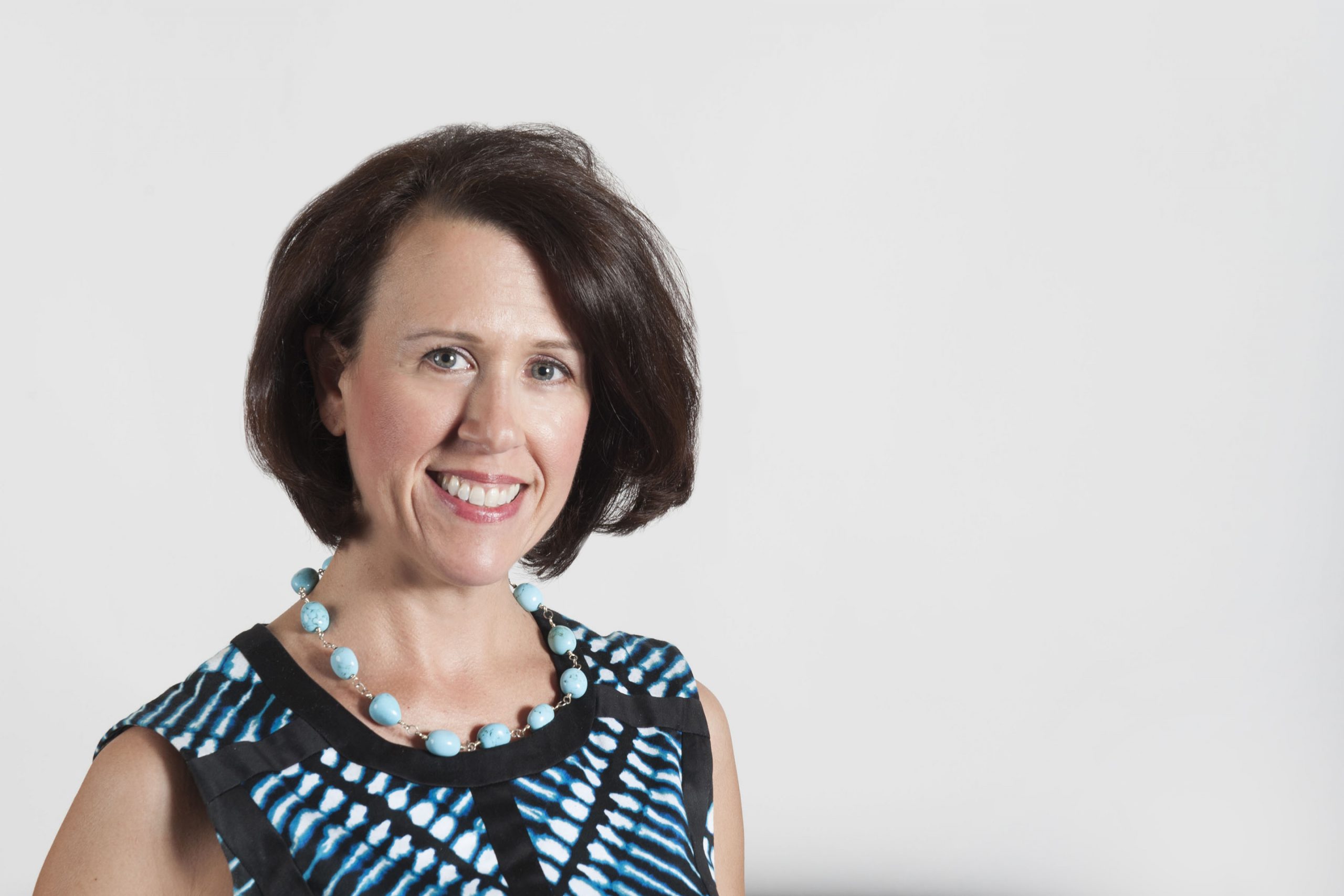 White woman with blue eyes, short brown hair with a big smile. She is wearing a blue neckless against a black and blue shirt.