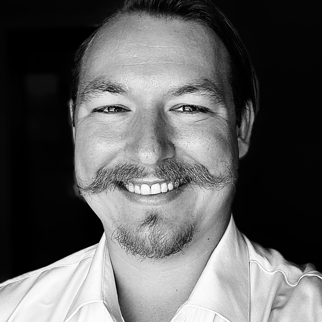 Black-and-white portrait of a white man with a pointed mustache, wearing a white collared shirt, facing forward with a neutral expression against a softly blurred background.