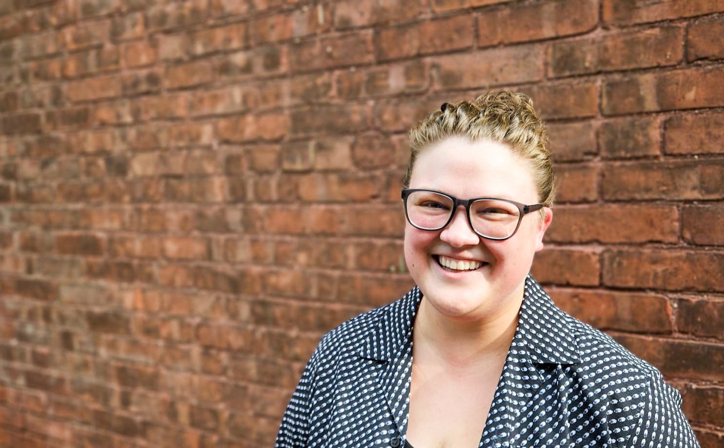 Blonde white female with black glasses on, bright smile and a collar shirt thats black and white
