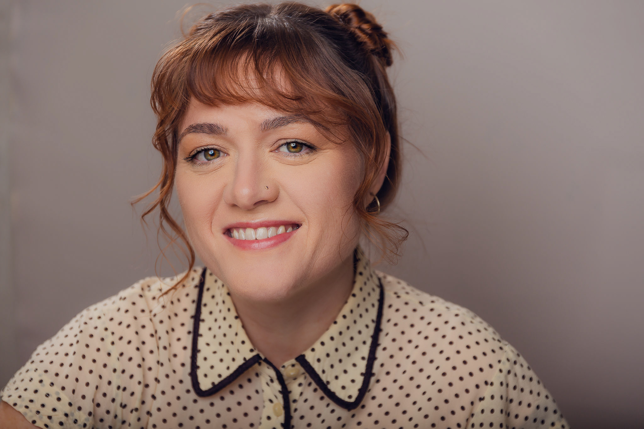 White woman with green/yellow eyes and red hair. She has a big smile and is wearing a yellow shirt with black polka dots.